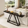 A dark wood console table with a modern geometric base is placed behind a white sofa in a well-lit living room. The table is decorated with a lamp, a bowl of yellow fruit, and a beige vase. A large window with a scenic view is in the background. Produktbild-Vorschau