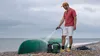A man is cleaning a green canoe on a pebble beach with a portable pressure washer. Anteprima dell’immagine prodotto