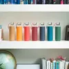 Seven glass water bottles with silicone sleeves in various colors are lined up on a white shelf. Produktbild-Vorschau