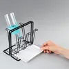 A hand is shown placing a drip tray under a black wire drying rack that holds a glass bottle with a light blue accessory. Náhled obrázku produktu