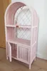 A pink wicker shelf unit with an arched top and two lower doors is shown in a room with a wooden door and a wall. Náhľad obrázku produktu