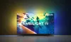 A Philips Ambilight TV is displayed in a dark room, with ambient lighting from the TV casting colors onto the wall behind it. Produktbild-Vorschau