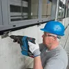 A man wearing a blue hard hat and safety glasses is using a Bosch cordless hammer drill to drill into a concrete wall. He is wearing work gloves and earplugs. Produktbild-Vorschau