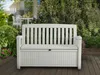 A white storage bench is placed on a wooden deck in a garden setting, surrounded by lush greenery and flowers. In the background, a white table and chairs are visible on a patio. Náhľad obrázku produktu