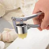 A hand is using a garlic press to crush garlic onto a white plate, with whole garlic bulbs in the background. Náhled obrázku produktu