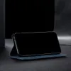 A blue flip-style phone case is shown standing upright on a dark surface, with a black smartphone inserted into it. The background is dark and out of focus. Produktbild-Vorschau
