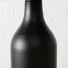 A close-up shot of a matte black object, possibly a lamp or vase, against a white brick wall. Produktbild-Vorschau