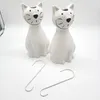 Two white ceramic cat-shaped humidifiers sit on a white surface with two S-hooks in front of them. Podgląd zdjęcia produktu