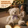 A young girl is sitting on the floor next to a large teddy bear, looking up at a decorated Christmas tree. Náhľad obrázku produktu