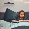 A young girl is reading a book in bed, with a shelf of books and fairy lights in the background. Produktbild-Vorschau