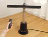 A hand holding a remote control is shown in the foreground, with a black and silver fan in the midground, set against a wooden floor and a white curtain in the background. Podgląd zdjęcia produktu