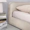A close-up view of a beige upholstered bed with a stack of books and a lamp on the left side. Produktbild-Vorschau