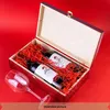 An open wooden wine box filled with red shreddings, containing two bottles of wine and a wine glass next to it, all on a red background. Produktbild-Vorschau