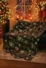 A Christmas-themed blanket with glowing patterns is draped over a sofa in a festive living room setting, with a decorated Christmas tree and stockings visible in the background. Náhled obrázku produktu