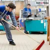 Two workers in a workshop setting. One is sweeping the floor with a broom, and the other is looking at a clipboard. Produktbild-Vorschau