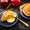A close-up of a slice of apple tart with custard on a dark plate, with whole apples and cinnamon sticks in the background. Produktbild-Vorschau