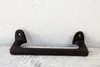 A rusty dark brown cast iron handle is laid horizontally on a light-colored wooden surface with visible grain against a textured white wall. Anteprima dell’immagine prodotto
