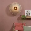 A wall lamp with a woven shade is mounted on a textured pink wall above a shelf with a picture and a plant. Below the shelf are two pillows. Anteprima dell’immagine prodotto