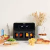 A black double air fryer is displayed on a white countertop, surrounded by various food items and kitchen accessories. Voir les images du produit