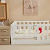 A white wooden crib with bedding and a red toy inside, placed next to storage boxes and a wall with a framed picture. Anteprima dell’immagine prodotto