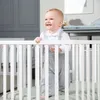 A baby is standing in a white crib, smiling and looking to the side. The background shows a nursery setting with gray furniture and a framed picture on the wall. Náhled obrázku produktu