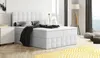 A white boxspring bed with storage is shown in a modern bedroom setting with a yellow rug and a floor lamp. Produktbild-Vorschau