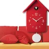 A red cuckoo clock with a pendulum hangs on a white background, with red pillows and a yellow couch visible in the foreground. Produktbild-Vorschau