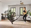 A woman is standing next to a stationary bike in a bedroom setting. Podgląd zdjęcia produktu