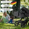 A woman and a child are sitting on the grass next to a black stroller in a park. Produktbild-Vorschau