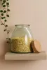 A glass storage jar filled with light yellow flakes, with a cork lid resting beside it, placed on a wooden shelf against a textured wall. A green vine hangs from the top left. Produktbild-Vorschau