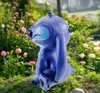 A blue Stitch figurine is sitting in a garden surrounded by flowers and greenery. Produktbild-Vorschau