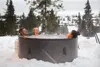 Three people relaxing in a hot tub outdoors in the snow, with trees and a fire pit in the background. Produktbild-Vorschau