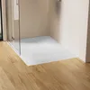 A modern bathroom corner with a white stone-look shower tray and a glass shower enclosure, set against a light tan wall and a wooden floor. Náhľad obrázku produktu
