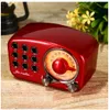 A vintage-style red radio with a dial and speaker grille sits on a wooden surface, with a blurred background of a lantern and plants. Produktbild-Vorschau