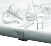Multiple clear plastic table clips are scattered on a white surface, with one clip attached to a table edge. Produktbild-Vorschau