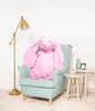 A large pink plush bunny sits in a light blue armchair next to a floor lamp and two small side tables. Náhled obrázku produktu
