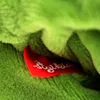 A close-up of a red tag with white text on a green plush toy. Produktbild-Vorschau