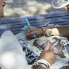 Two people are sitting on a bench and decorating a piece of clothing with markers and rhinestones. Voir les images du produit