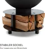 A close-up view of a black metal fire pit base with stacked firewood stored underneath. Produktbild-Vorschau