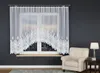 A white lace curtain with a decorative pattern is hung over a window, with a city skyline visible through the glass. A cream-colored armchair and a small side table with a lamp and books are placed to the right of the window. Náhled obrázku produktu