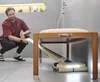 A man crouches down looking at a cordless floor cleaner working under a wooden bench with a white cushion. Anteprima dell’immagine prodotto