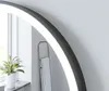 A close-up view of a round mirror with integrated LED lighting, reflecting a plant and a towel rack. Produktbild-Vorschau