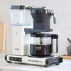 A white Moccacoffee maker with a glass carafe filled with coffee sits on a kitchen counter. Produktbild-Vorschau