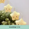 A string of cute, star-shaped LED lights with smiling faces, set against a blurred background of greenery and a plain wall. Náhľad obrázku produktu