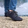 A person wearing blue jeans and dark blue shoes stands on a rocky surface next to a body of water. Produktbild-Vorschau