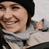 A close-up of a woman wearing a black beanie and a gray scarf, smiling. Náhled obrázku produktu