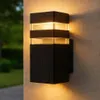 A black outdoor wall light mounted on a textured concrete wall, with a blurred green garden in the background. Náhľad obrázku produktu
