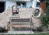 A rustic garden bench sits in front of a brick wall with a decorative window and a wagon wheel. Produktbild-Vorschau