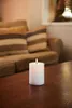 A white LED candle with a lit flame sits on a wooden table in front of a blurred couch. Produktbild-Vorschau