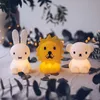 Three small, illuminated animal-shaped night lights (a rabbit, a lion, and a bear) are arranged on a textured surface, possibly a tablecloth, with blurred greenery and fairy lights in the background. Anteprima dell’immagine prodotto